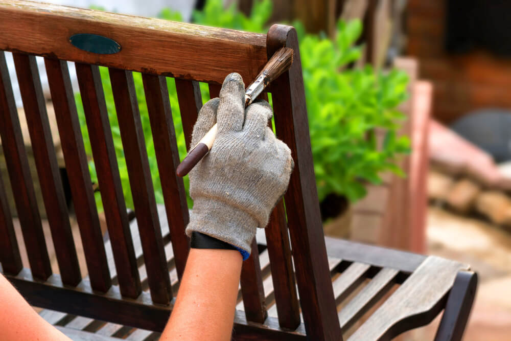 A hand paints stain onto a wooden bench as part of their patio furniture restoration process